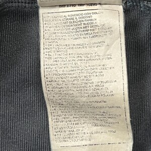 May include: Close-up of a dark gray fabric tag with printed text in multiple languages. The tag is attached to a dark gray garment. The text includes care instructions and a product code.