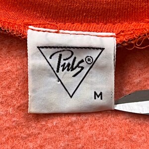 May include: Close-up of a white clothing tag with the brand name "Puls" inside a black triangle. The tag is attached to an orange fabric. The size "M" is printed below the logo.