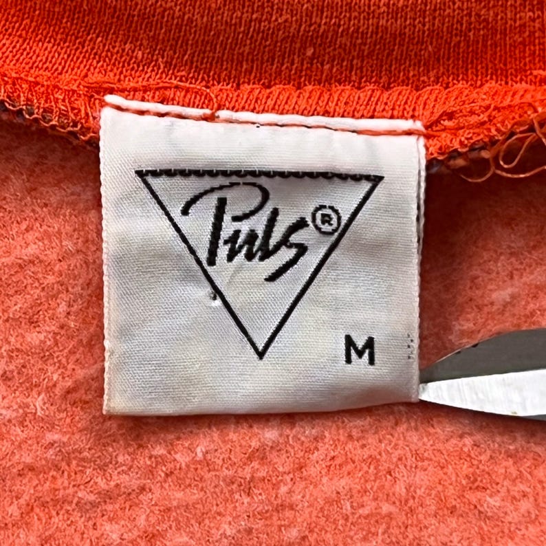 May include: Close-up of a white clothing tag with the brand name "Puls" inside a black triangle. The tag is attached to an orange fabric. The size "M" is printed below the logo.