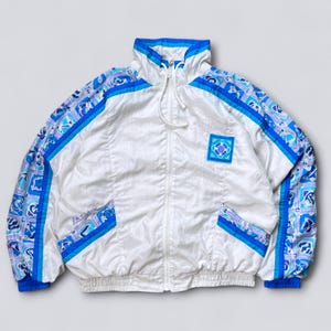 May include: White vintage track jacket with blue and purple patterned accents on the sleeves and shoulders. Features a high collar, a full zipper, and a square patch on the chest. The jacket has blue trim and pockets.