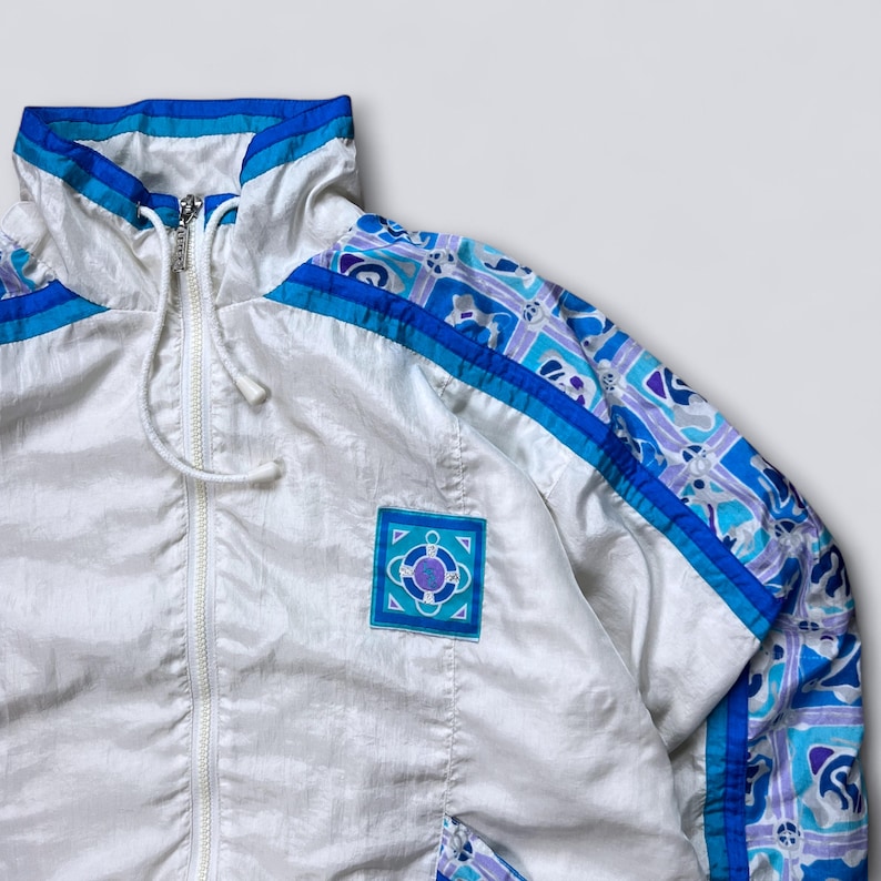 May include: White vintage track jacket with blue and purple accents. The jacket features a high collar, a full zipper, and a drawstring at the hem. The sleeves have a patterned design in blue, purple, and white. A square patch with a geometric design is on the chest.