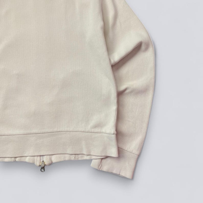 以下が含まれることがあります： リブ編みのテクスチャが特徴の、ライトベージュのジップアップスウェットシャツ。ゴールドカラーのジッパーと、リブ編みの裾と袖口が付いています。画像右側に袖が見えます。