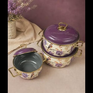May include: A set of three round enamel pots with matching lids, featuring a cream-colored base with purple floral patterns and small bird illustrations. Each pot has gold-toned metal handles and a solid purple lid with a gold-toned knob handle.