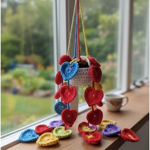 May include: A handmade crochet hanging planter with a white basket and colorful heart accents. The planter is suspended by blue, red, and yellow cords. Red, blue, yellow, green, and purple crochet hearts cascade from the basket, creating a whimsical, decorative piece.
