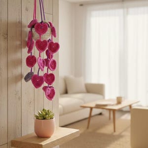 Puede incluir: Una maceta colgante de ganchillo hecha a mano con decoraciones en forma de corazón rosa. La maceta está suspendida de un cordón morado y rosa. Una pequeña planta suculenta en una maceta rosa se encuentra en un estante de madera.