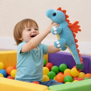 Op de afbeelding: Een gehaakte blauwe draak met oranje accenten, witte stippen en een gele buik. De draak wordt vastgehouden door een kind in een ballenbak vol kleurrijke ballen. De draak heeft grote zwarte ogen en een lange staart.