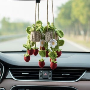 Op de afbeelding: Twee hangende gehaakte aardbeienplant auto-ornamenten. Elk ornament heeft een beige pot, groene bladeren, rode aardbeien en witte bloemen. De ornamenten zijn opgehangen aan beige koorden.