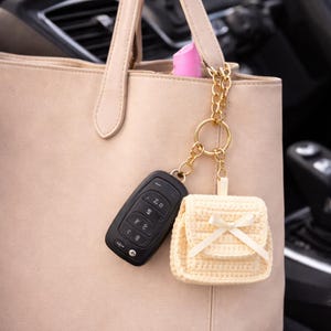 Puede incluir: Un bolso tote de cuero beige con una cadena dorada y un pequeño llavero de mochila de ganchillo. Una llave de coche negra está unida al llavero. La mochila llavero tiene un lazo color crema.