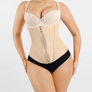 Peut inclure: Un corset beige avec une fermeture éclair sur le devant et des bretelles réglables. Le corset est porté sur un soutien-gorge beige et un sous-vêtement noir. Le vêtement est conçu pour façonner le torse.
