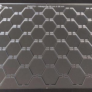 Peut inclure: Plaque en acrylique transparente avec un motif en nid d'abeille de découpes hexagonales. La plaque est étiquetée "PM02T01 - Hexágones 26 mm X 25 mm" et comporte de petits trous dans chaque coin. Le site web www.sellagab.com est visible.