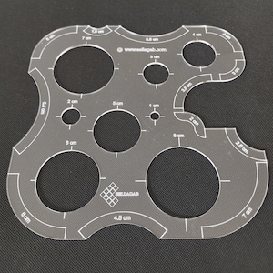 Könnte beinhalten: Transparente Acryl-Quiltvorlage mit verschiedenen kreisförmigen Aussparungen und gebogenen Kanten. Die Vorlage weist Messungen in Zentimetern von 0,5 cm bis 8,5 cm sowie die Website-Adresse www.sellagab.com auf.