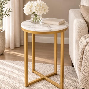 May include: A round side table with a white faux marble top featuring grey veining and a gold-finished metal frame with an X-shaped base. A clear glass vase filled with white hydrangea flowers and a closed beige book sit on the tabletop surface.