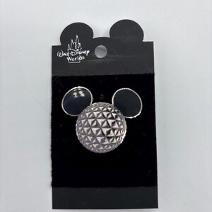 May include: A pin featuring a geometric sphere design with a purple and silver finish, flanked by two black circles, resembling Mickey Mouse ears. The pin is attached to a black backing with the "Walt Disney World" logo.