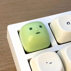 Peut inclure: Une touche de clavier artisanale de forme carrée, de couleur vert clair, arborant un visage minimaliste avec deux yeux ronds sombres, un petit nez triangulaire et une bouche en ligne droite, installée sur un clavier mécanique blanc.