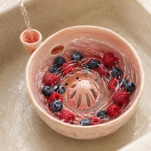 May include: A light pink ceramic berry colander bowl with a small side handle and a central drainage dome. The bowl is filled with fresh blueberries and raspberries being rinsed under a stream of water, showing clear water flowing through the holes.