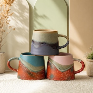 Puede incluir: Tres tazas de cerámica apiladas, cada una con una combinación de colores única. La taza superior es azul, la del medio es turquesa y la inferior es rosa. Cada taza tiene un asa y un acabado texturizado y moteado.