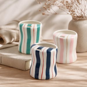 May include: Three ceramic vases with vertical stripes in teal, pink, and navy blue with red and gold accents. Each vase has a unique, curved shape and a small opening at the top. A neutral-toned vase with dried flowers is in the background.