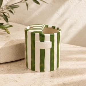 Può includere: Un vaso in ceramica a righe verdi e bianche, con apertura a forma di cuore. Ha una piccola etichetta bianca con la scritta "coffee time". Il vaso cilindrico è posto su una superficie chiara. Ideale per fiori o decorazioni.