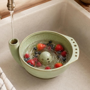 May include: A light green ceramic berry colander with a central perforated dome and a side spout for water intake. The bowl is filled with water, fresh red strawberries, and dark blue blueberries, with a faucet stream flowing into the spout.