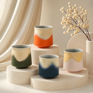 May include: Four ceramic tumblers in various colors: orange, green, blue, and pink. Each tumbler has a unique two-tone design with a light-colored top and a darker base. The tumblers are displayed on textured, off-white pedestals.