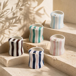 May include: Five ceramic vases with vertical striped patterns in various colors: brown and white, teal and red, blue and red, pink and gold, and light blue. Each vase has a unique design and a small opening at the top. The vases are arranged on a stone surface.