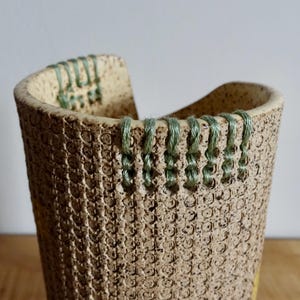 May include: A cylindrical ceramic vessel with a speckled beige surface featuring a textured grid pattern. The upper rim is accented with sage green thread stitched through small holes, creating a decorative border around the top edge of the object.