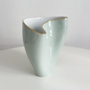 May include: A pale green ceramic vase with a unique, asymmetrical shape. The rim is outlined with a thin gold band. The interior is white, creating a contrast with the exterior. The vase is a decorative item.