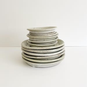 May include: A stack of ceramic plates in varying sizes, featuring a light gray glaze with dark brown speckles throughout. The plates have a slightly raised rim and a smooth, rounded edge, creating a uniform appearance in this set of stoneware.