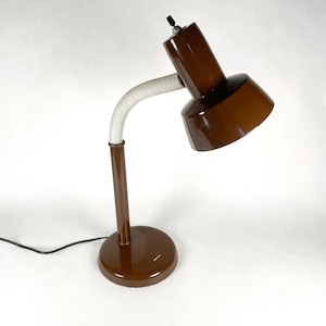 May include: A brown desk lamp with a round base, a vertical stem, and a flexible, white, ribbed neck. The lamp head is angled and has a black on/off switch. The lamp is approximately 12 inches tall.