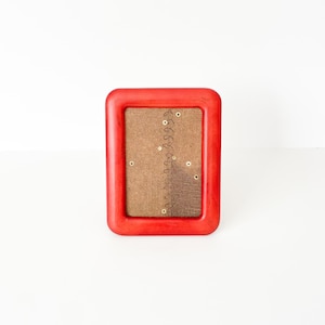 May include: A rectangular picture frame with a thick, smooth, bright red border. The center insert is made of a textured, tan-colored burlap fabric featuring a vertical line of small, dark brown embroidered stitches and several small, circular metal eyelets.