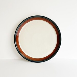 May include: A round ceramic plate featuring a smooth, cream-colored center basin. The plate is framed by a wide, deep brown rim that transitions into a thin, dark black edge, creating a distinct contrast between the light interior and dark border.