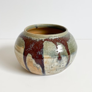 May include: A round ceramic vase with a wide opening. The vase has a speckled, light-colored exterior with dark brown drips and a green rim. The interior is a warm, earthy tone.