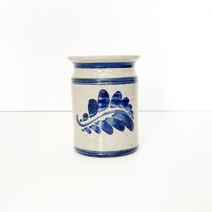 May include: A cylindrical stoneware crock featuring a speckled light gray finish with dark blue horizontal bands at the top and bottom. The center of the vessel is decorated with a hand-painted dark blue leaf motif consisting of several segments.