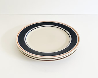 May include: A stack of three round ceramic plates featuring a cream-colored center and a wide dark navy blue rim. The rim is accented by thin, concentric orange and navy blue circular lines that frame the edge of each plate in the set.