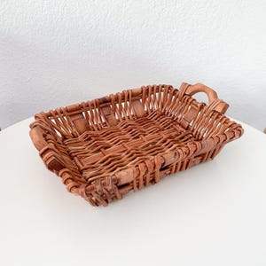 May include: A rectangular woven wicker basket with a handle on each end. The basket is made of light brown wicker and has a woven pattern. The handles are curved and made of the same material. The basket is empty.