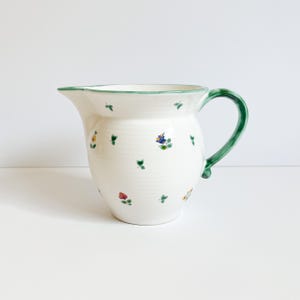 May include: A white ceramic pitcher featuring a hand-painted pattern of small, colorful flowers and green leaves scattered across the body. The pitcher has a curved handle and a green painted rim, with a spout for pouring liquids.