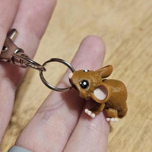 Puede incluir: Un llavero de ratón marrón con un llavero y un cierre plateados. El ratón tiene ojos negros grandes, orejas blancas y pequeñas patas blancas. El llavero lo sostiene una persona.
