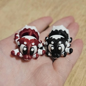 Puede incluir: Dos figuras de mofeta impresas en 3D. Una es roja y blanca, la otra es negra y blanca. Ambas tienen ojos grandes y de dibujos animados y están tumbadas sobre sus vientres. Las mofetas son pequeñas y parecen juguetes u objetos decorativos.