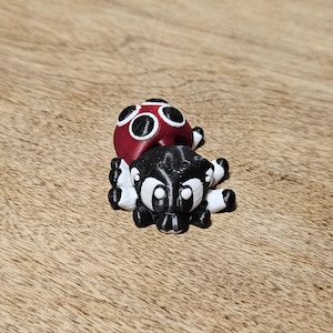 Puede incluir: Una criatura de juguete impresa en 3D con una cabeza roja y un cuerpo negro. La cabeza tiene detalles circulares blancos y el cuerpo tiene patas a rayas blancas y negras. El juguete está sobre una superficie de madera.