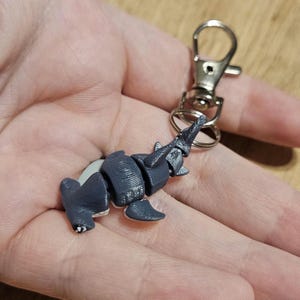May include: A gray elephant-shaped keychain charm with a silver-colored clasp. The elephant is segmented and has detailed texture. The keychain is held in a hand against a neutral background.