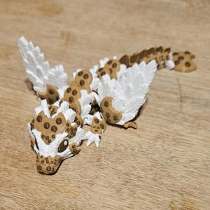 May include: A whimsical, 3D-printed dragon figurine with a unique cookie-themed design. The dragon features white wings and body accents, with brown cookie details, including chocolate chips. The figurine is posed on a wooden surface.
