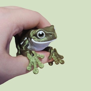 May include: A detailed, metallic green frog figurine with white accents. The frog has large, expressive eyes and is posed with its front legs extended. The figurine is held in a hand, showcasing its size and detail. The frog's skin has a textured appearance.