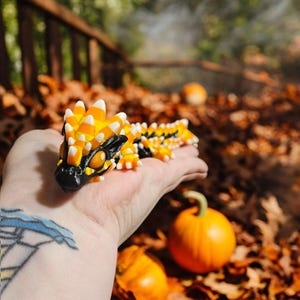 Puede incluir: Figura detallada de un dragón con cabeza negra, cuerpo amarillo y detalles blancos y naranjas. El dragón descansa sobre una mano, con calabazas y hojas de otoño en el fondo. Un artículo decorativo único.