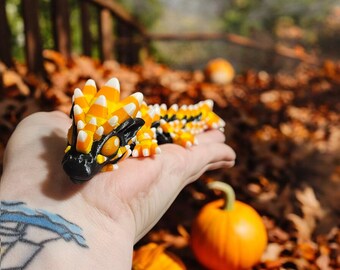 May include: A detailed, colorful dragon figurine with a black head, yellow and orange body, and white accents. The dragon is resting on a hand, with a blurred background of fall foliage and pumpkins. The figurine is a unique, handcrafted item.