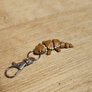 May include: A brown polymer clay keyring shaped like a segmented creature, possibly a caterpillar or worm. The keyring has a silver-coloured clasp and ring. The creature has white accents and small black eyes. The background is a wooden surface.