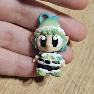 May include: A miniature elf figurine with a green and white hat, green hair, and a green and white outfit. The elf has large black eyes and a black belt. The figurine is held in a hand.