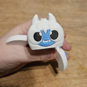 以下が含まれることがあります： 大きな黒い目、青い口、小さな角を持つ、白と青の3Dプリントされた漫画のモンスターのフィギュア。 フィギュアには翼があり、手で持たれています。