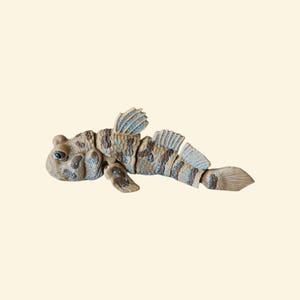 May include: A detailed, segmented fish figurine in shades of brown, tan, and blue. The fish has a rounded head, detailed fins, and a segmented body with a pattern of spots and stripes. The figurine is set against a neutral background.