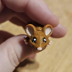 Puede incluir: Una pequeña figura de ratón marrón impresa en 3D con grandes orejas con bordes blancos, ojos negros y una nariz blanca. El ratón se sostiene en una mano, mostrando su diseño y textura detallados.