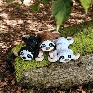 Pode incluir: Três estatuetas de bicho-preguiça em preto, castanho e branco com garras e traços faciais castanhos repousam sobre um tronco coberto de musgo. As preguiças têm olhos grandes e expressivos e estão deitadas. O fundo inclui folhas e um cenário natural desfocado.